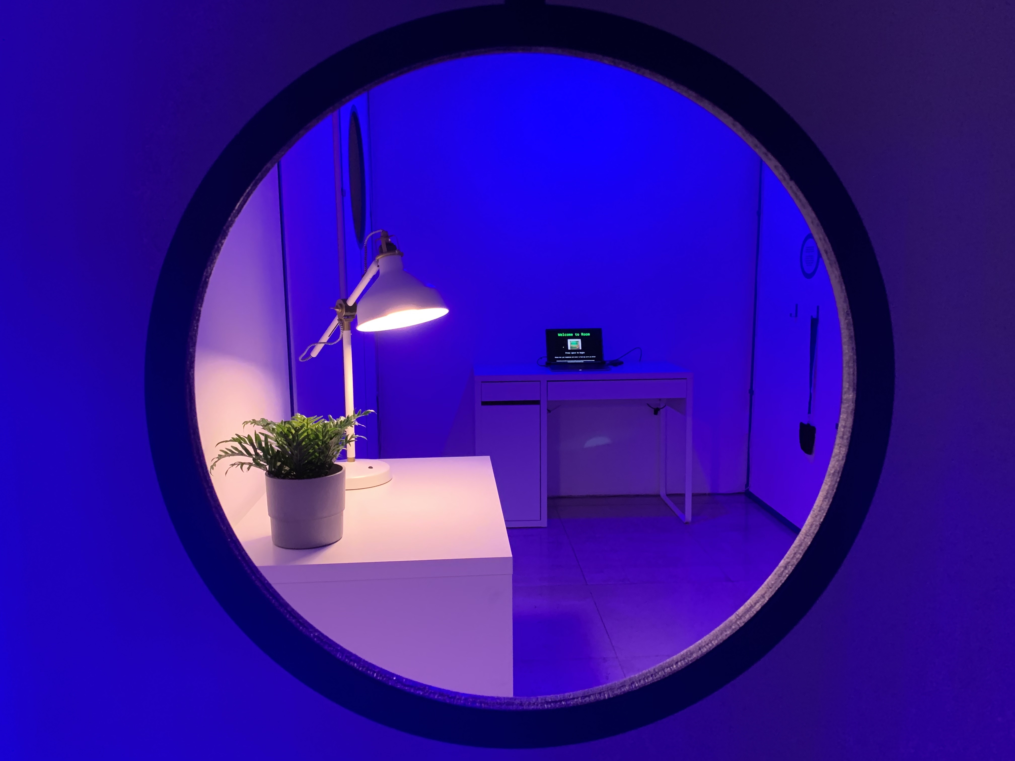 A round window looking into a room bathed in purple light with a white desk, lamp and plant on top, 